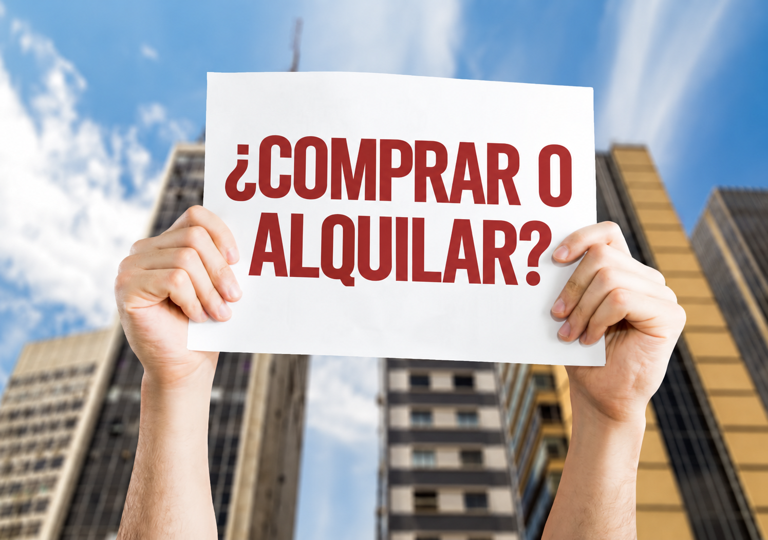 Manos sosteniendo un cartel con el texto «¿Comprar o alquilar?» frente a edificios.